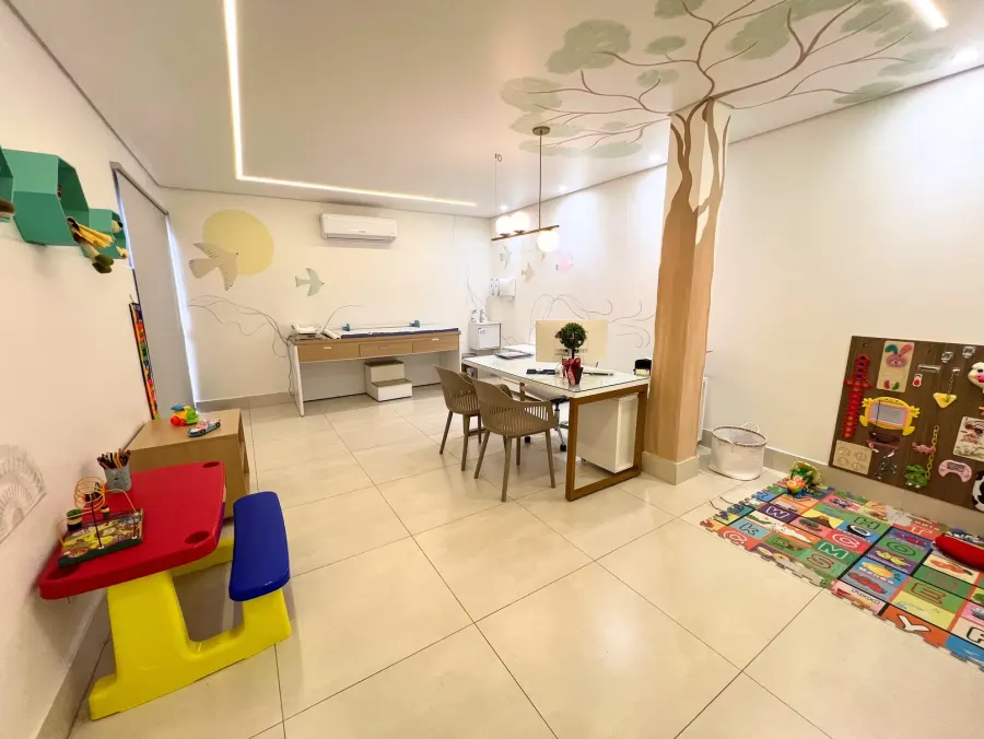 Consultório de pediatria da Viva Pediatria: mesa de atendimento, maca de exame, mesinha de brincar e painel de atividades, com uma árvore pintada na parede.