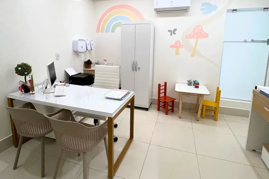 Mesa de atendimento com duas cadeiras para os pais, armário e mesinha infantil ao fundo, em consultório de pediatria.