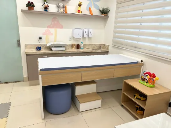 Maca de exame pediátrico com balança de bebê sobre a bancada, ao lado de um móvel com brinquedos.