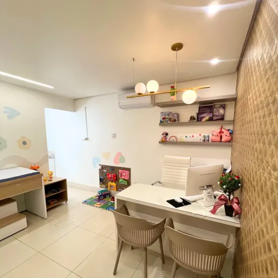 Mesa de atendimento com duas cadeiras para os pais, prateleira com livros de pediatria e painel de atividades para as crianças.