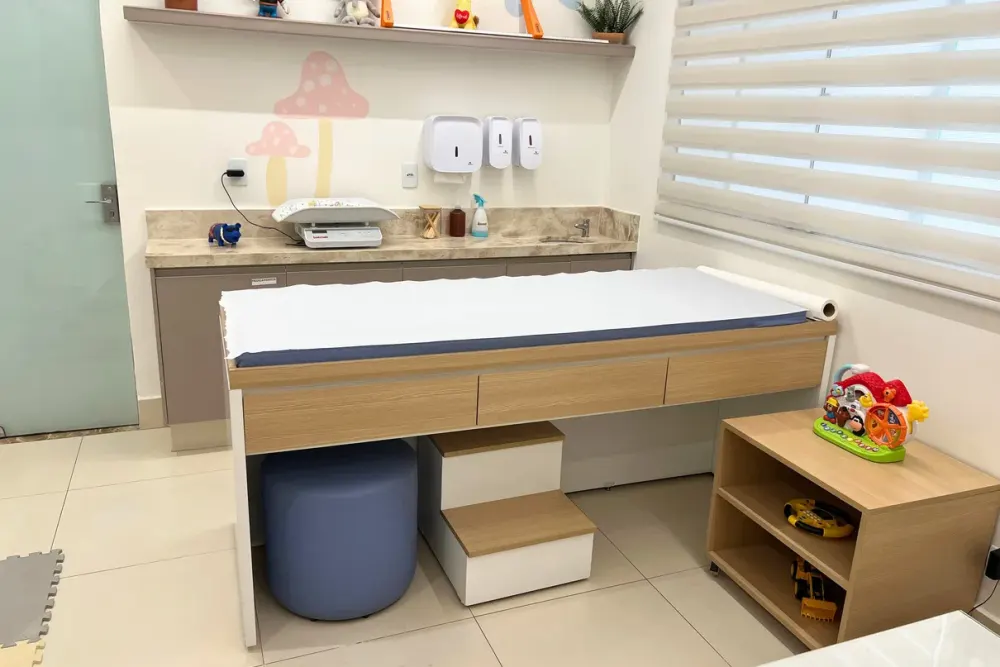 Maca de exame pediátrico com balança de bebê sobre a bancada, ao lado de um móvel com brinquedos.