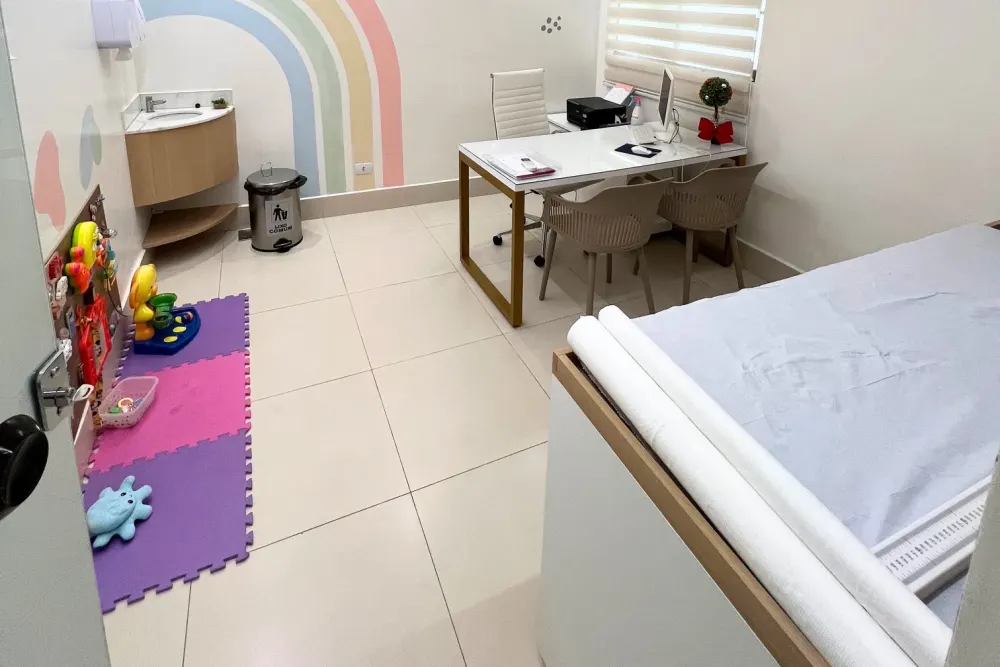 Consultório de pediatria da Viva Pediatria: maca de exame, pia, tapete de brinquedos e mesa de atendimento com cadeiras para os pais.