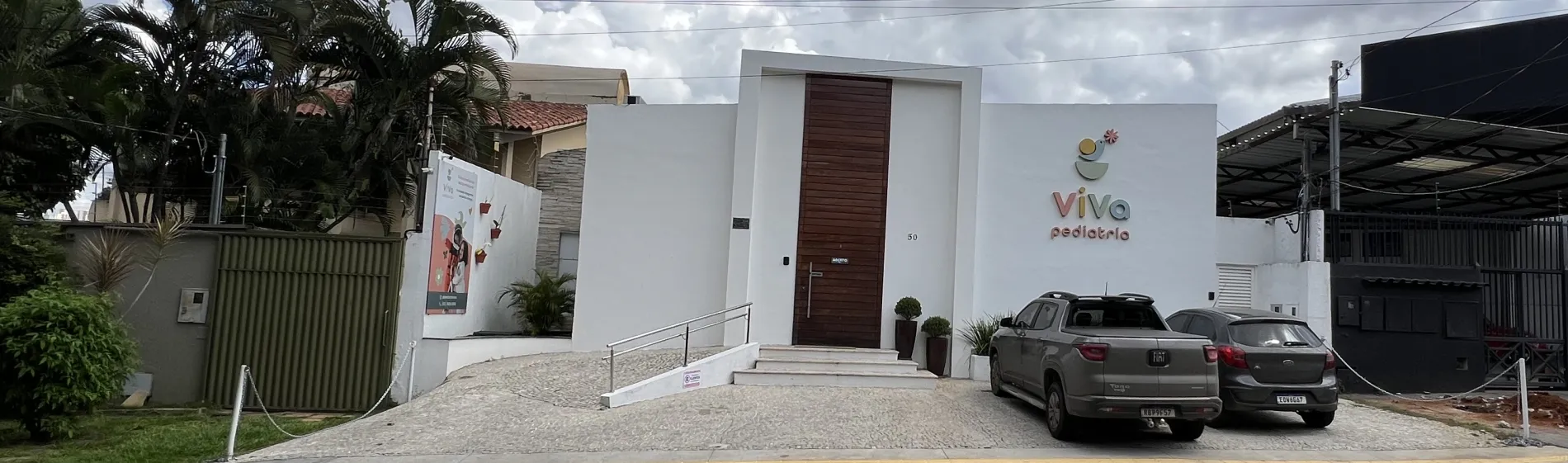 Fachada da Viva Pediatria vista da rua: prédio branco com porta de madeira, o letreiro da clínica na parede e vaga de estacionamento em frente.