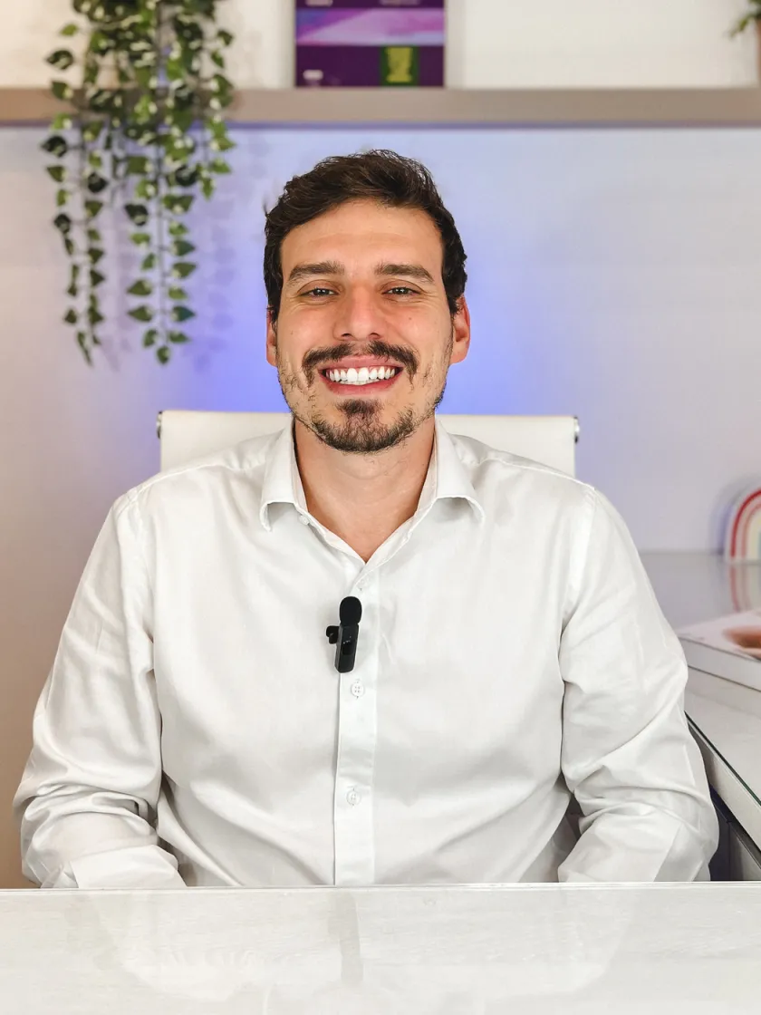 Dr. Pedro Conelian em seu consultório, de camisa branca, sentado à mesa de atendimento.